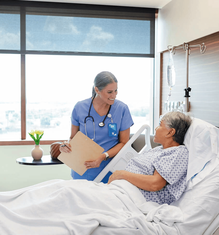 Smiling nurse in blue scrubs talking to an elderly female patient in a hospital bed during daytime, with a clipboard in hand and an IV stand beside the bed in a well-lit, modern hospital room.